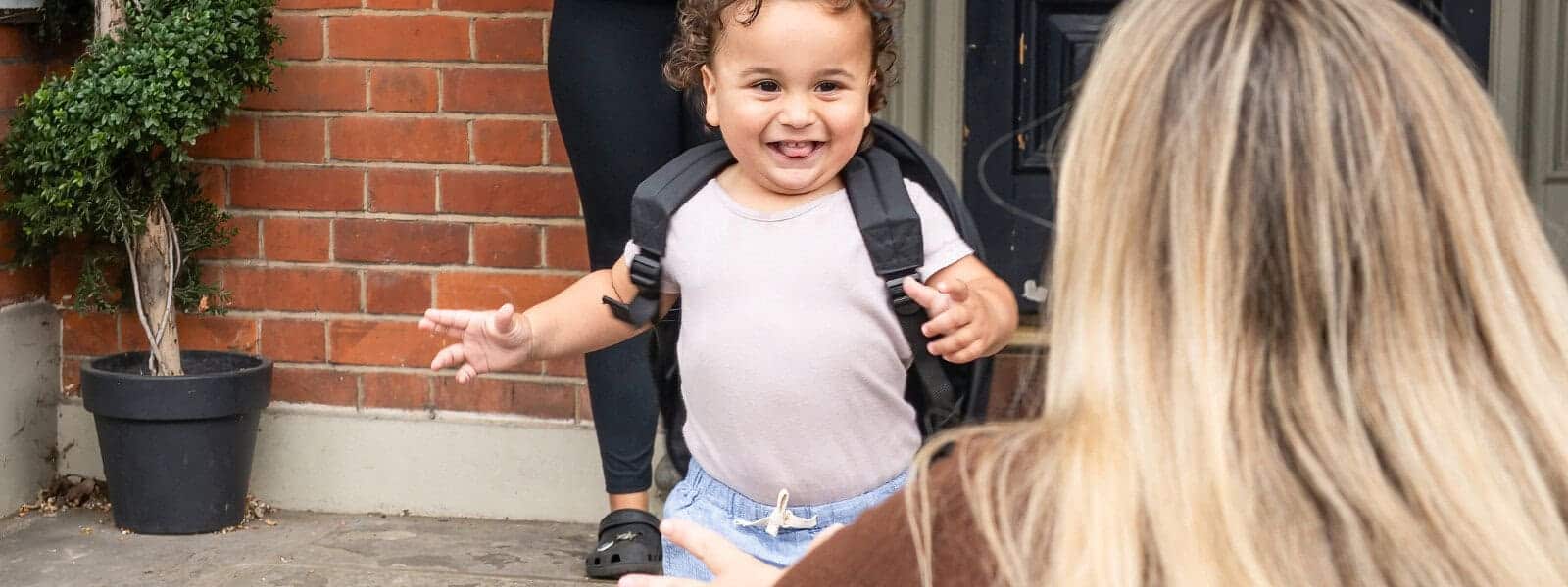 Colorful toddler with backpack smiling outdoors, preparing for nursery at Monkey Puzzle Day Nurseries, warm welcome for young children, engaging early childhood education.