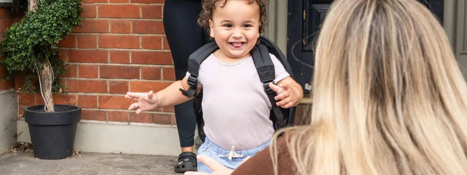 Colorful toddler with backpack smiling outdoors, preparing for nursery at Monkey Puzzle Day Nurseries, warm welcome for young children, engaging early childhood education.